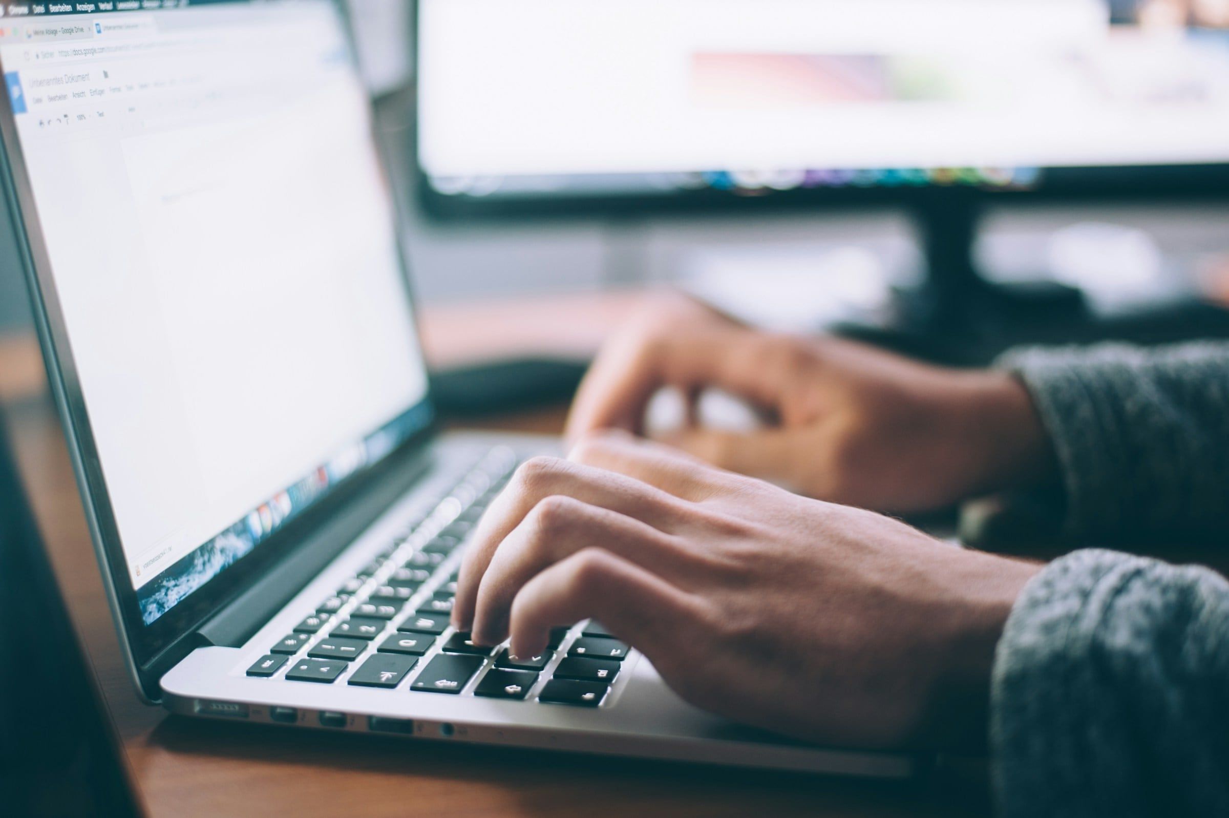 Manos escribiendo en una laptop frente a una pantalla con documentos de consulta